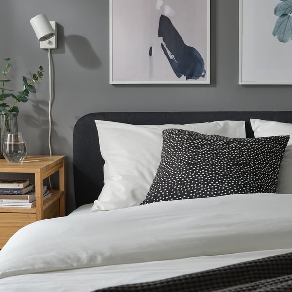 Cosy grey bed with white & dotted cushions, wall art, small plant, wooden bedside table with books.