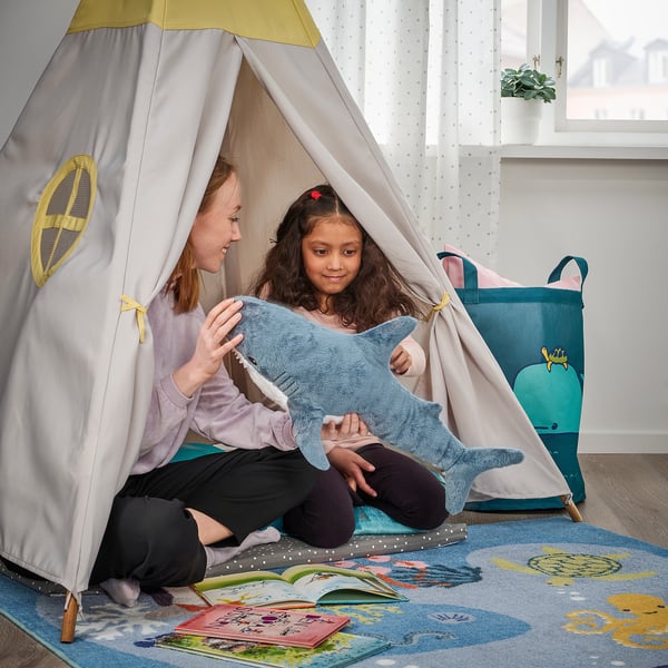 Two kids play inside a tent with a soft shark toy. Books and a storage bin are nearby.