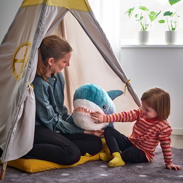 Child in tent with adult and shark toy. Adult sits, child reaches for toy. Grey tent, green plants nearby.