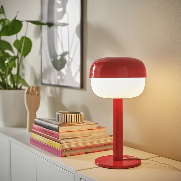 Red BLÅSVERK lamp on desk, modern retro design, glossy finish, soft lighting, near books and plant.