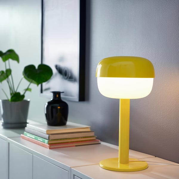 Bright yellow BLÅSVERK lamp on table, surrounded by books, plant, and vase; retro, soft shape, modern design, small size.