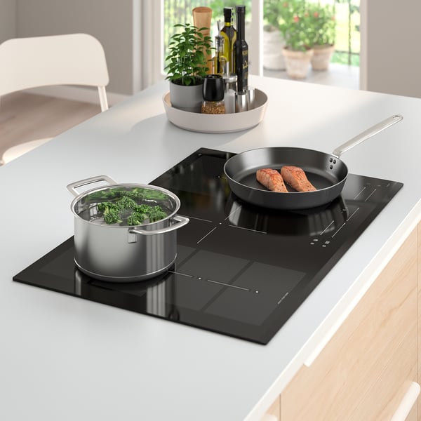 Modern kitchen with BLIXTSNABB induction hob, five zones cooking vegetables and sausages. Tray with bottles and plant beside.