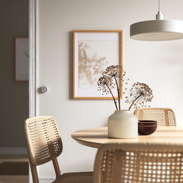 Minimalist dining area with round table, wicker chairs, vase, bowl, pendant lamp, nature print.