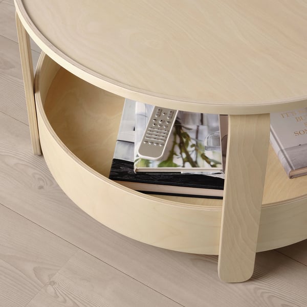 Light wood round coffee table with storage shelf, holding remote and books.