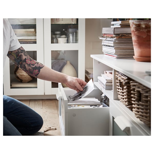 Hand with tattooed arm reaches into a white BRIMNES tv unit drawer, organising items. Beside the unit is a stack of books and a clay pot.