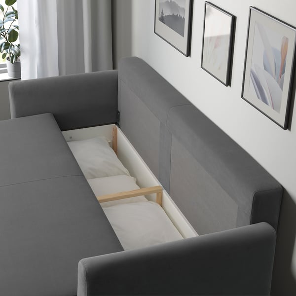 Modern grey sofa bed with storage, open to reveal white bed linens. Artwork on wall, plant nearby.