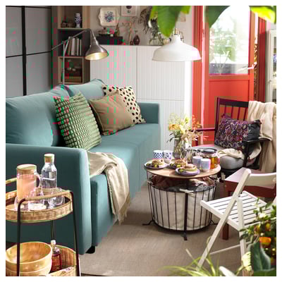 Cosy living room with teal sofa, colourful pillows, food-filled table, and decor. Bright light, red door.