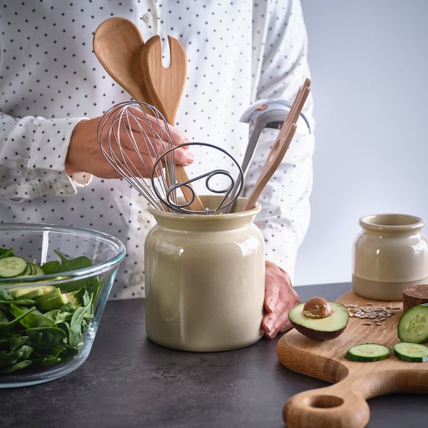 Person holding utensils in a stoneware holder on a kitchen counter with fresh salad and ingredients nearby.