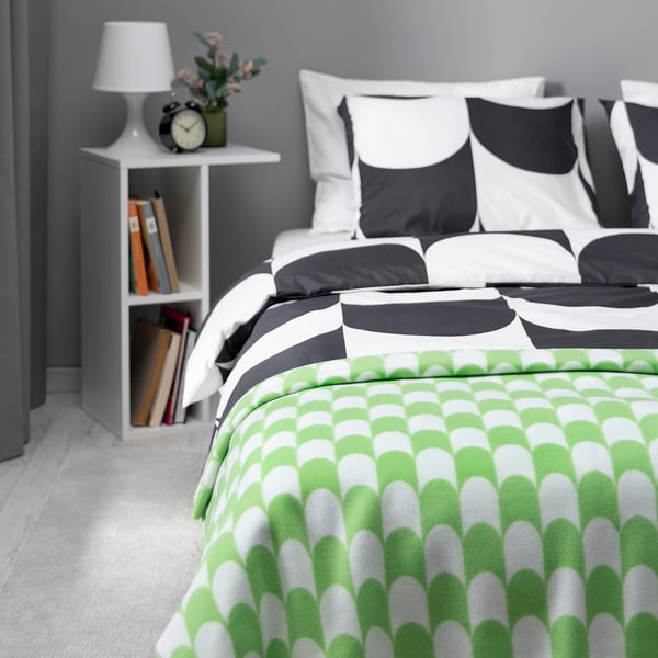 Modern bedroom with BRUKSVARA green throw blanket on bed, matching cushions, grey pillows, white lamp on small table with books.