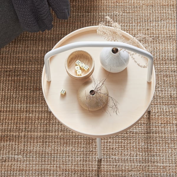Small round birch side table with handle, holding dice set, small vases, dried plants, set on woven beige rug.