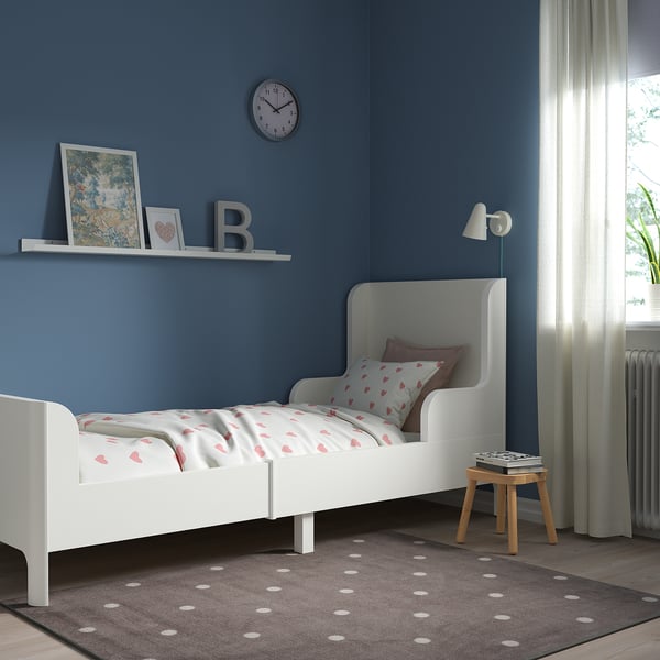 White kids bed in blue room. Heart bedding, stool beside, under window.