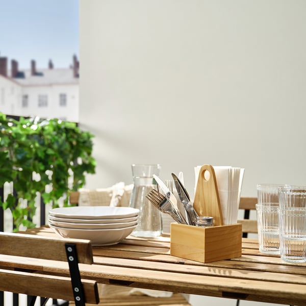 Wooden table holds two stacked bowls, clear glasses, and a CHOKLADHAJ organizer with cutlery and napkins, near green plant. Classic, rustic design.