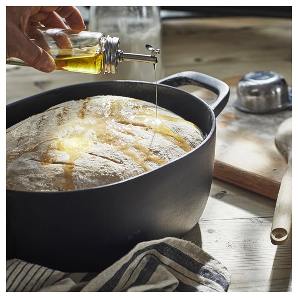 A kitchen scene a glass oil bottle being poured over dough in a black bowl. A striped towel rests beside it.