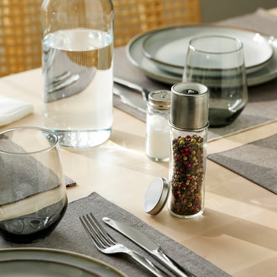 Table setting with clear glass spice grinder, plates, cutlery, and glasses on striped placemats.