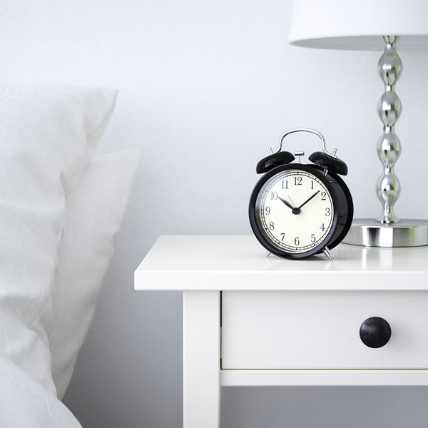 Black alarm clock on white bedside table, displaying time 12:30, beside a white pillow and lamp.
