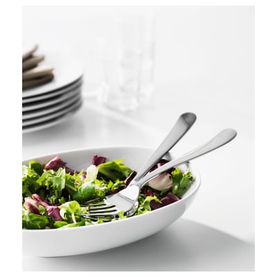 White bowl with green salad, two stainless steel forks resting on it, stack of plates and glass in background.