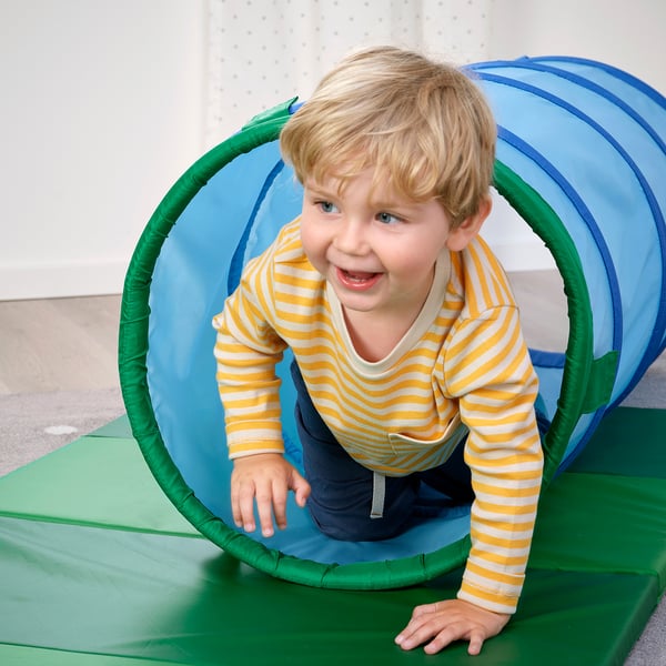 A child plays inside a colourful plastic tunnel, crawling through it on a green mat.