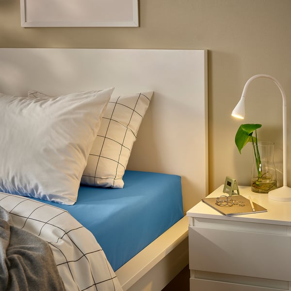 Modern bedroom: white bed with blue DVALA cotton sheet, white pillows, bedside table with lamp, plant, and jewellery.