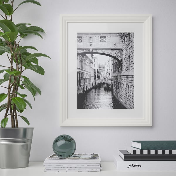 Framed venice bridge hangs on white wall. Beside it, a potted plant and book stack.