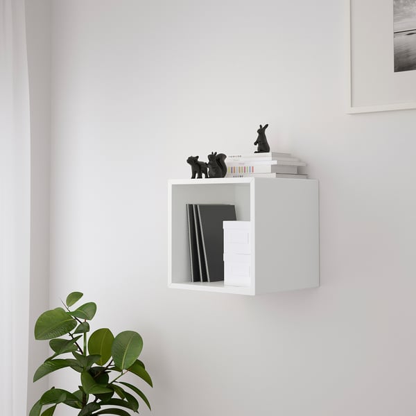 White wall-mounted storage cube holding books and black decorative figures, with a potted plant below.