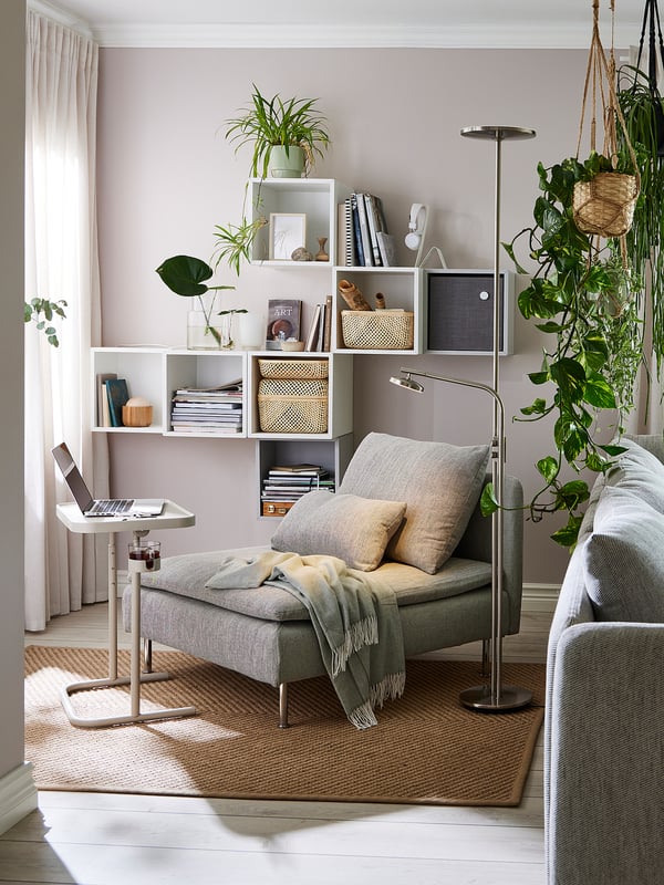 Modern living room with EKET shelving unit, grey couch, laptop table, green blanket, striped cushions, floor lamp, books, and hanging plants.