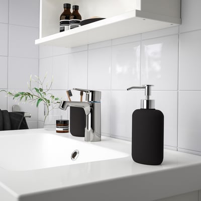 Modern bathroom sink with sleek chrome tap and black soap dispenser, small plant, toothbrush holder, and jars on counter, white tiles.
