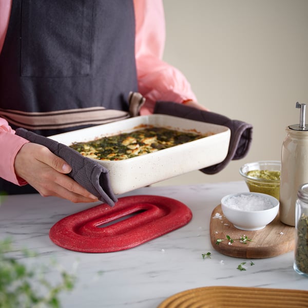 Person holding dish with baked food, using heat-resistant potholder on red bamboo trivet.