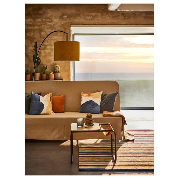 Cosy living room with tan sofa, colourful pillows, striped rug, lamp, and cacti near large windows showcasing a sunset.