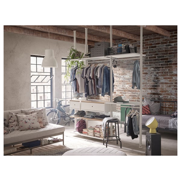 Modern loft-like room with exposed brick, a white sofa, and an open storage system. Clothes and books displayed on adjustable shelves and rails.