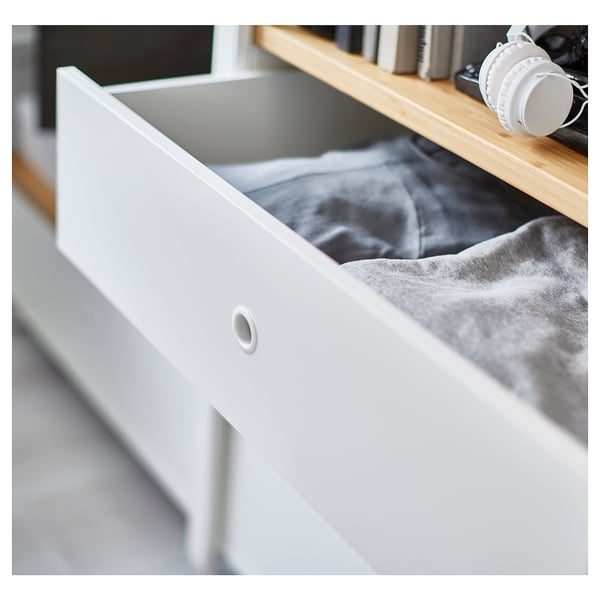 Open storage drawer in white metal and bamboo, partially filled with folded grey fabric.