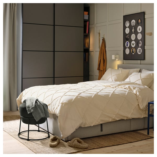 Bedroom with large bed, white blanket, grey headboard, grey chair, rug, slippers, and wall art.