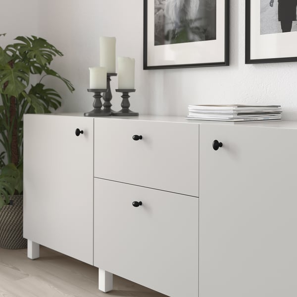 White cabinet with black knobs, holding candles and books, next to a plant.