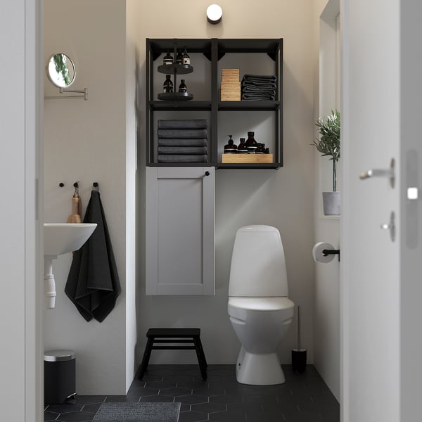 Modern bathroom with grey ENHET cabinet, white toilet, sink, and black accents. Storage shelves hold towels and toiletries.
