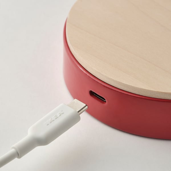 A red desk lamp with a wooden top and a usb-c port for charging a device is shown. A white usb-c cable is plugged into the lamp.