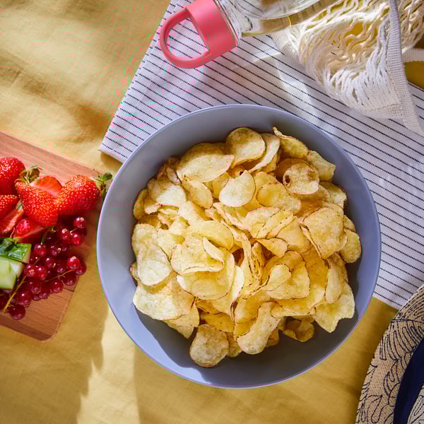 A bowl of ENSTÄMMIG blue potato chips on a yellow table with a fruit platter and a red pitcher.