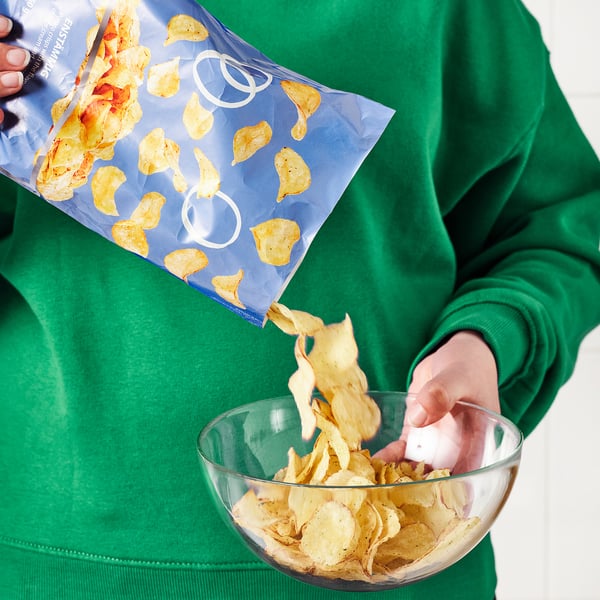 Person pouring potato chips into a glass bowl from a blue packet labelled ENSTÄMMIG while wearing a green sweatshirt.