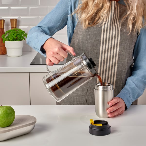 A person pours coffee from a clear pitcher into a stainless steel ENVÄLDIG travel mug with a black lid on a kitchen counter.