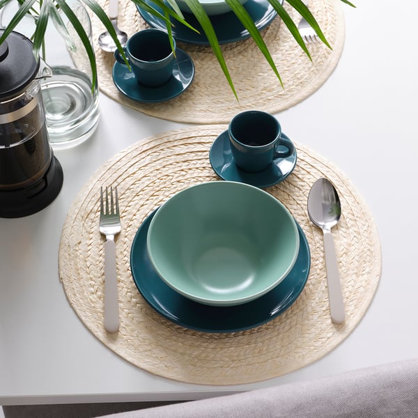 A table setting with green FÄRGKLAR bowls, cups, and woven placemats. Includes cutlery and a coffee press.