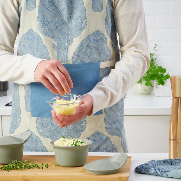Person wearing an apron adds grated cheese to a sauce in a dish. Dish is placed on a wooden cutting board with other kitchen items.