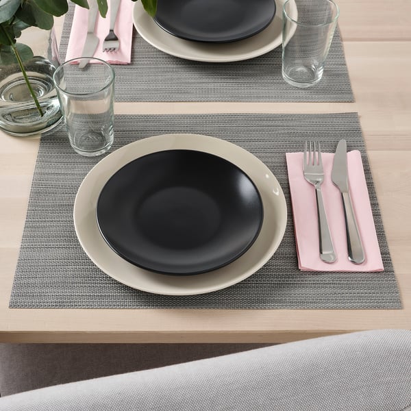 Two place settings on a table with grey placemats, black and white plates, pink napkins, forks, knives, glasses, and a small candle.
