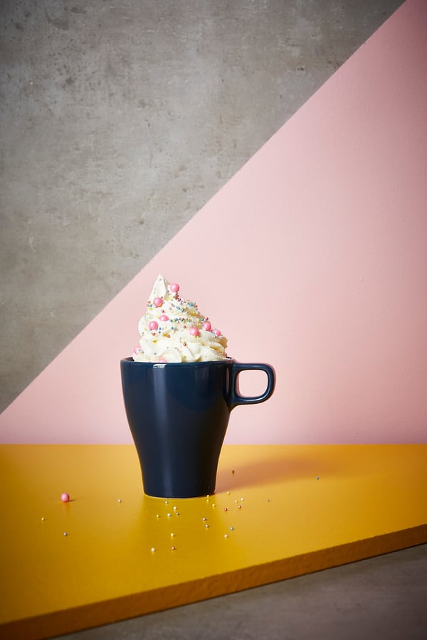 Dark blue mug with white whipped cream and colourful sprinkles on a yellow shelf against pink and grey walls.