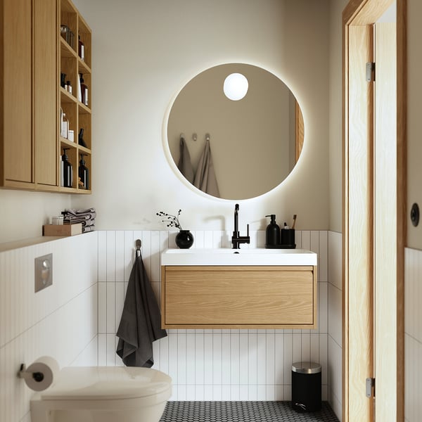 Modern bathroom with large round mirror, wooden vanity, and flowers.