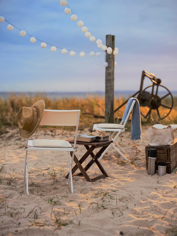 Beach setup with FEJAN white folding chairs, string lights, sand, hat, table, and wheelbarrow.