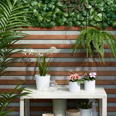 White FEJKA potted plant on a table, surrounded by other plants and flowers, set against a wooden wall.