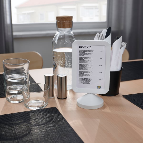 A café table set with a menu board displaying text in a frame, glasses, cutlery, napkins, and a water carafe.