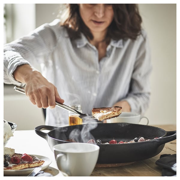 A person using a stainless steel FINMAT spatula, holding a cooked food slice over a skillet with berries, on a wooden table with a cup of coffee.