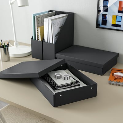 A black FJÄDERHARV storage box on a desk, holding papers and notebooks, near a lamp and art prints.