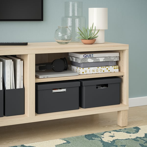 Light wood storage shelf with black boxes, books, lamp, plant, and headphones, great for organising and decor.