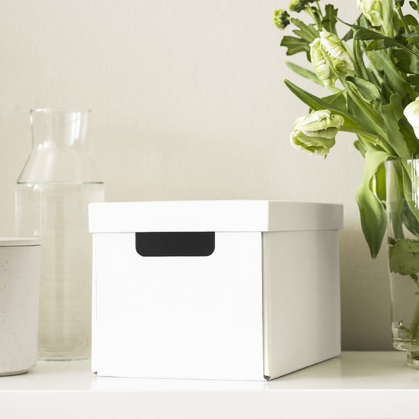 White box with black handle sits on white table near glass vase and green flowers. Ideal for storing small items.