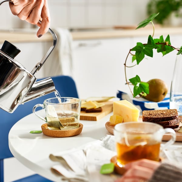 Hand pours tea from kettle into clear glass mug on coaster, surrounded by cheese, crackers, and another mug of tea on a table.